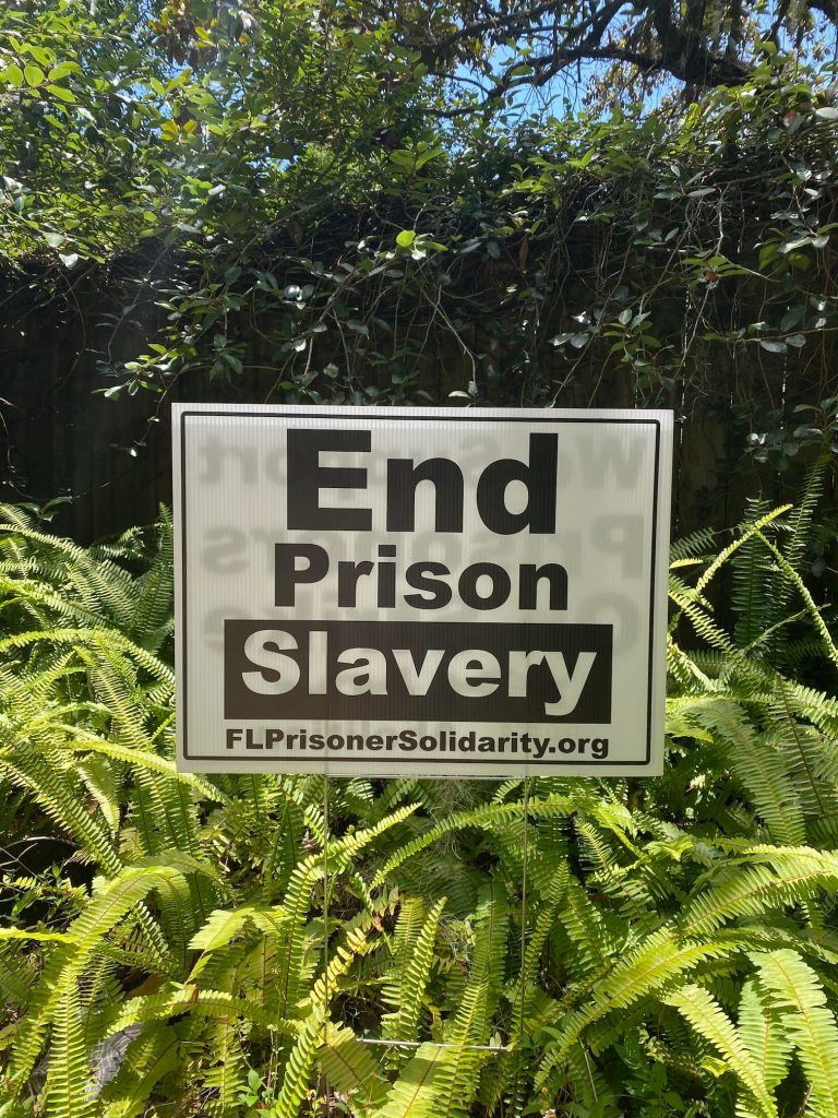 A yard sign within ferns that reads "End prison slavery" with the URL for flprisonersolidarity.org.